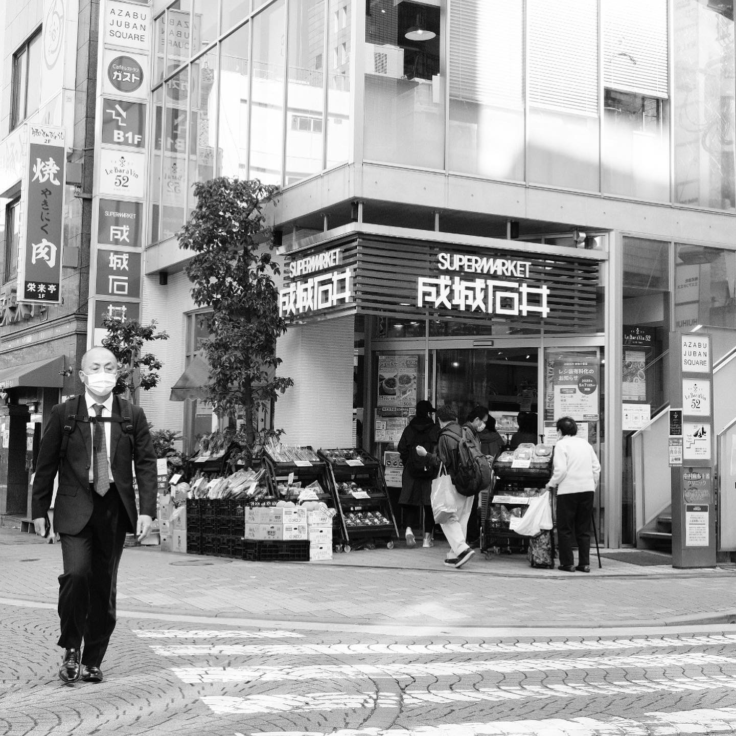 Black and white photograph of a bustling street scene outside a supermarket in Azabu Juban, Tokyo. A businessman in a suit and face mask walks across a patterned sidewalk, while shoppers enter and exit the store. Fresh produce is displayed outside the entrance, and various signs in Japanese and English, including ‘Supermarket’ and ‘Azabu Juban Square,’ are visible on the surrounding buildings.