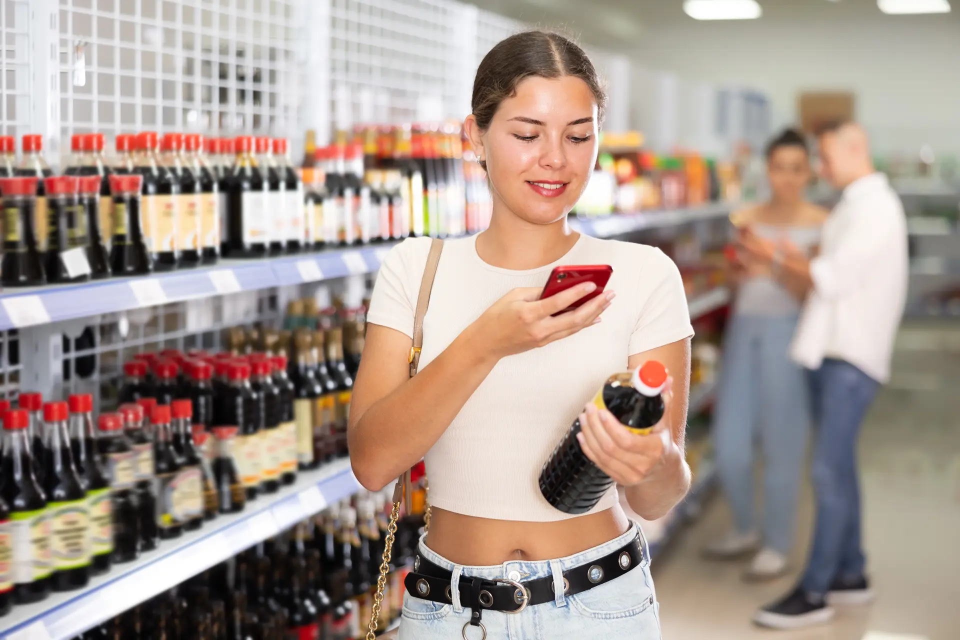 A young woman in a grocery store aisle scanning a bottle of soy sauce with her phone, likely researching ingredients or comparing products. Shelves stocked with various bottles of soy sauce and condiments line the background.