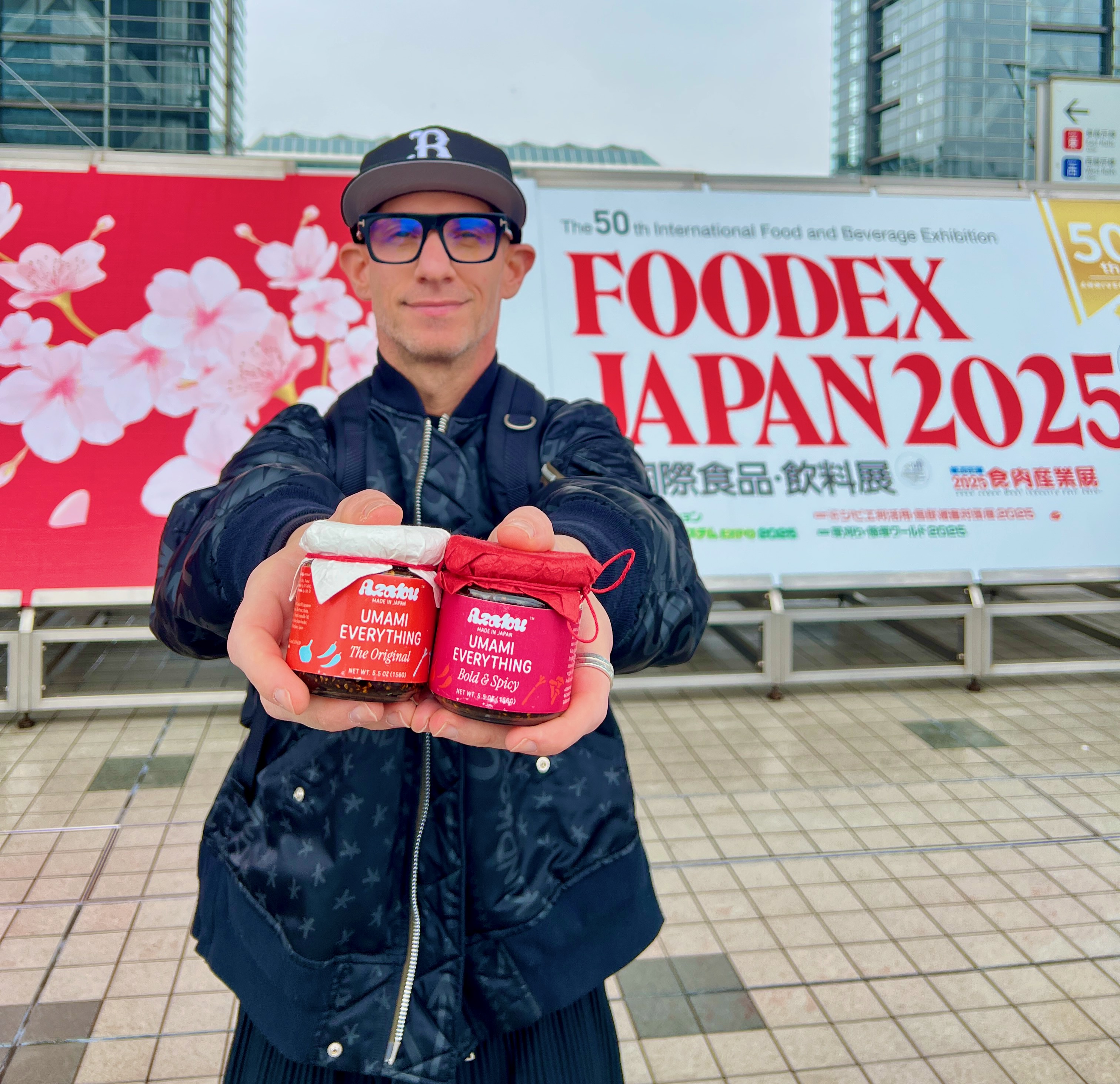 Brendan, founder of Azabu Foods, holding jars of Umami Everything – The Original and Bold & Spicy – in front of the FOODEX Japan 2025 exhibition in Tokyo.
