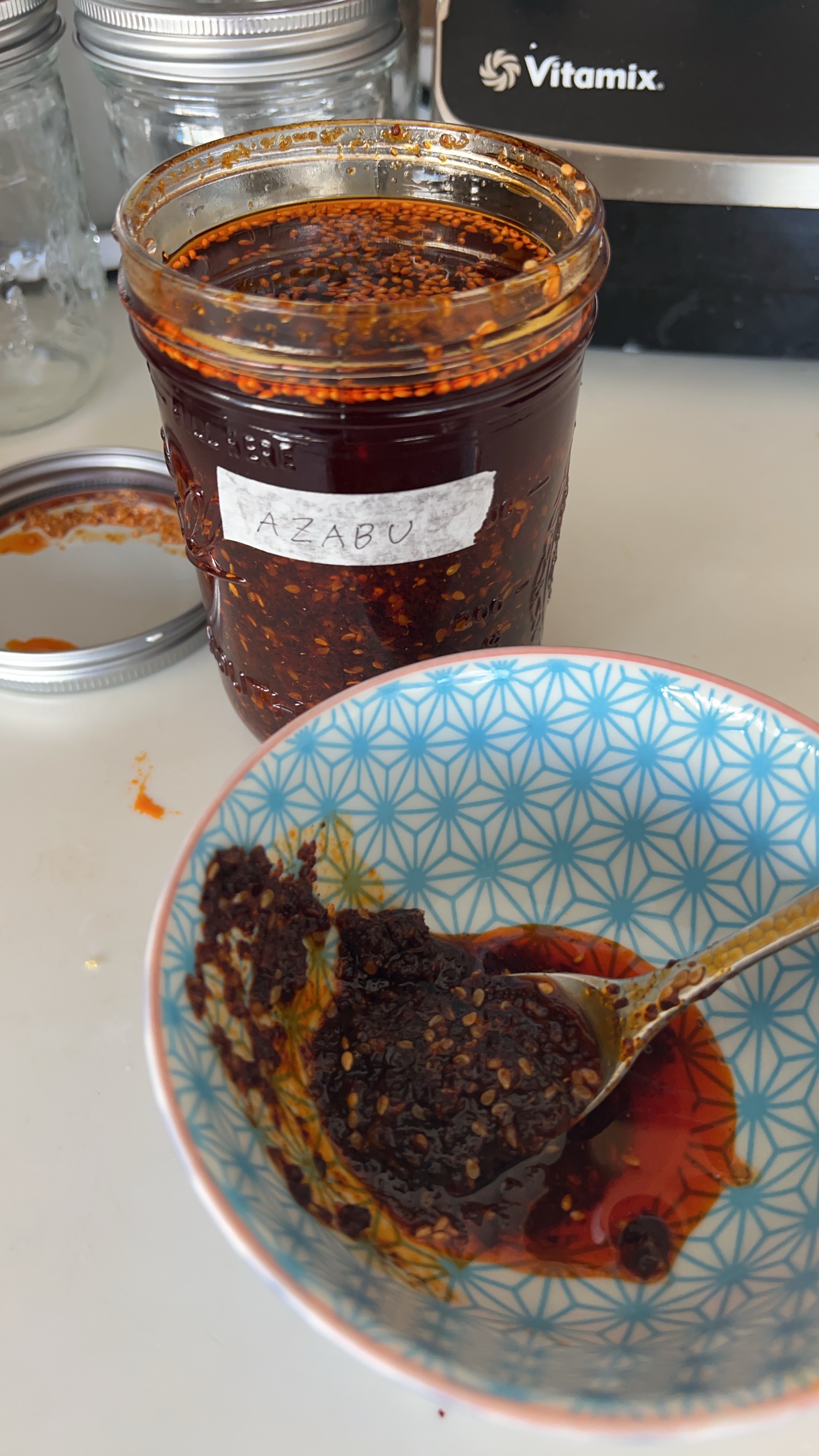 A jar of homemade chili oil labeled “AZABU” sits open on a kitchen counter, with a spoonful of the rich, umami-packed mixture scooped into a patterned blue and white bowl. The deep red oil glistens with sesame seeds and chili flakes, showcasing its bold flavor. A Vitamix blender and glass jars are visible in the background, suggesting a test kitchen setting for Azabu Foods’ Umami Everything.